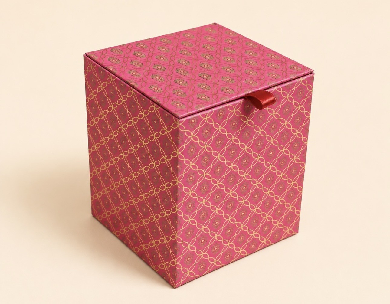 Closed pink cube gift box with a gold-coloured trellis pattern and a red ribbon pull tab.